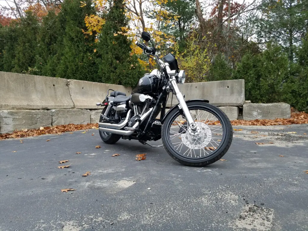 Black Harley-Davidson Dyna motorcycle parked on asphalt with concrete wall and autumn trees in background.