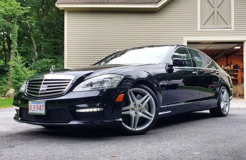 Black Mercedes-Benz sedan parked in driveway in front of garage with wooded background.