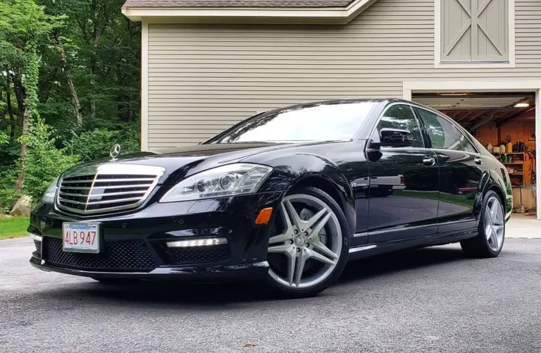 Black Mercedes-Benz sedan parked in driveway in front of garage with wooded background.