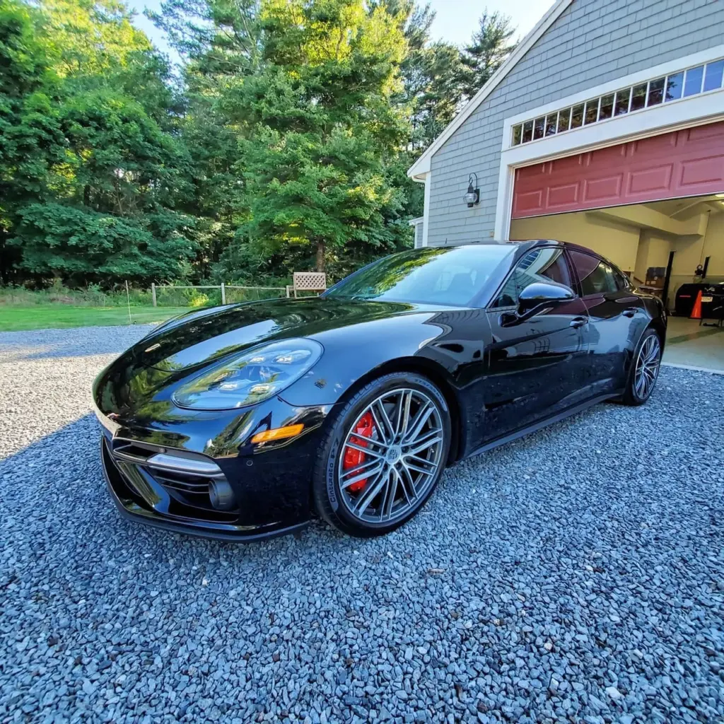 Black Porsche 911 sports car parked on gravel driveway in front of garage with red door.