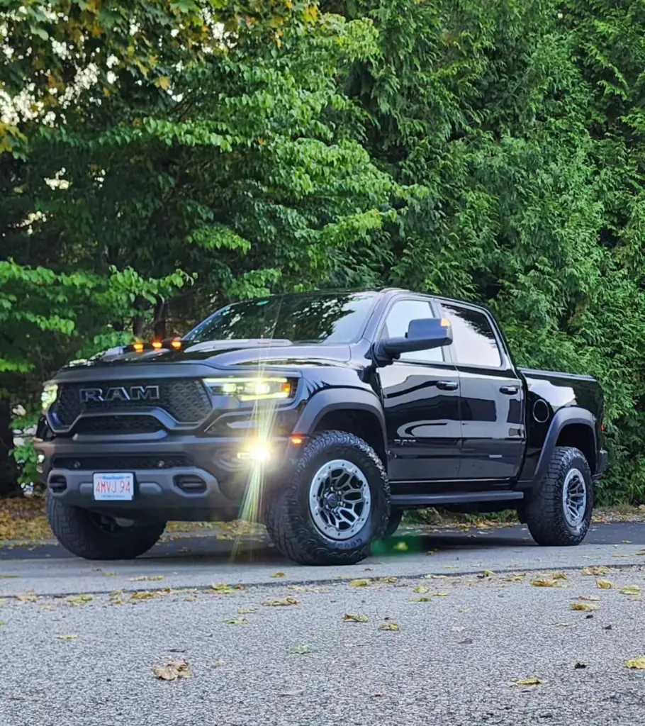 Black RAM pickup truck with illuminated headlights parked on driveway with green foliage backdrop.