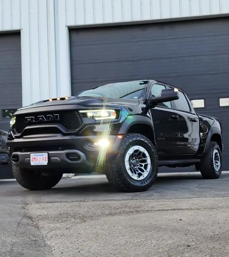 Black RAM pickup truck with illuminated headlights parked in front of a detailing facility garage.
