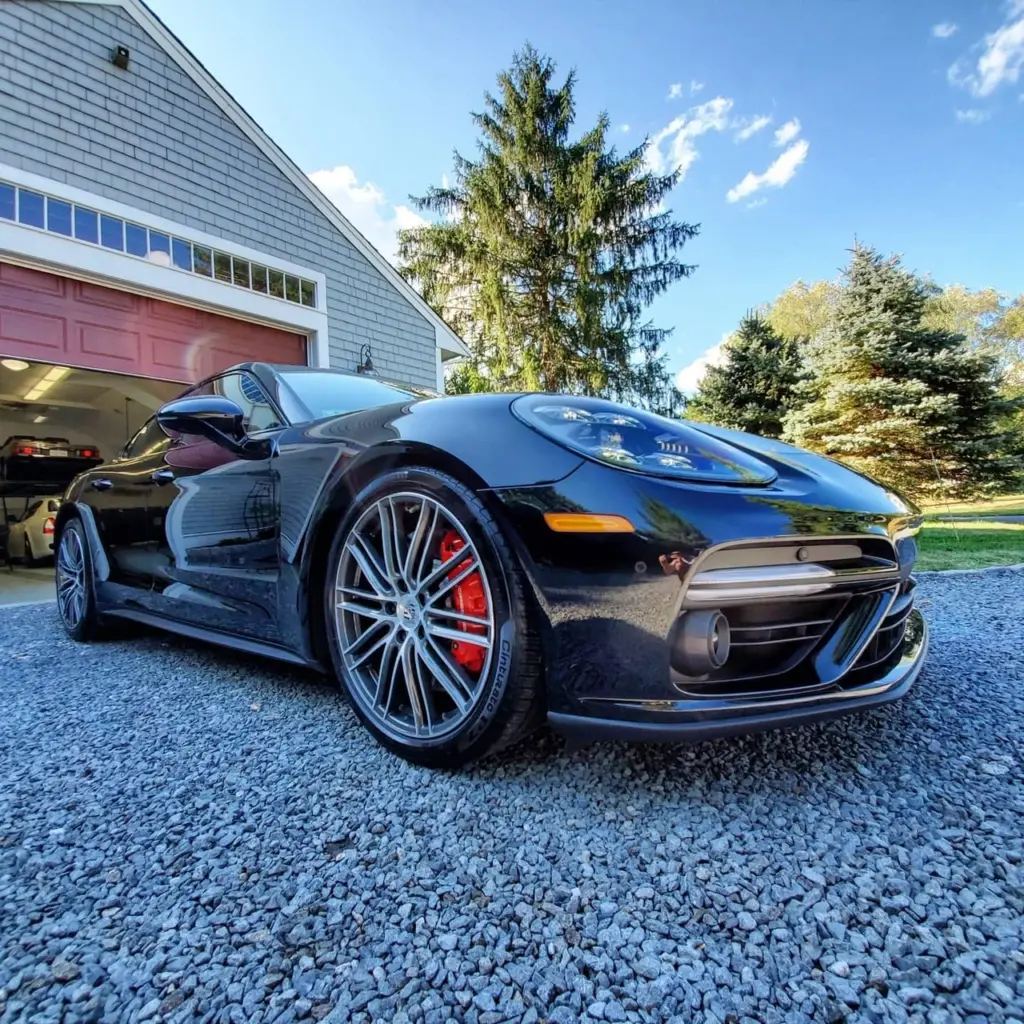 Black sports car with open driver door parked on gravel driveway beside garage with pink door.