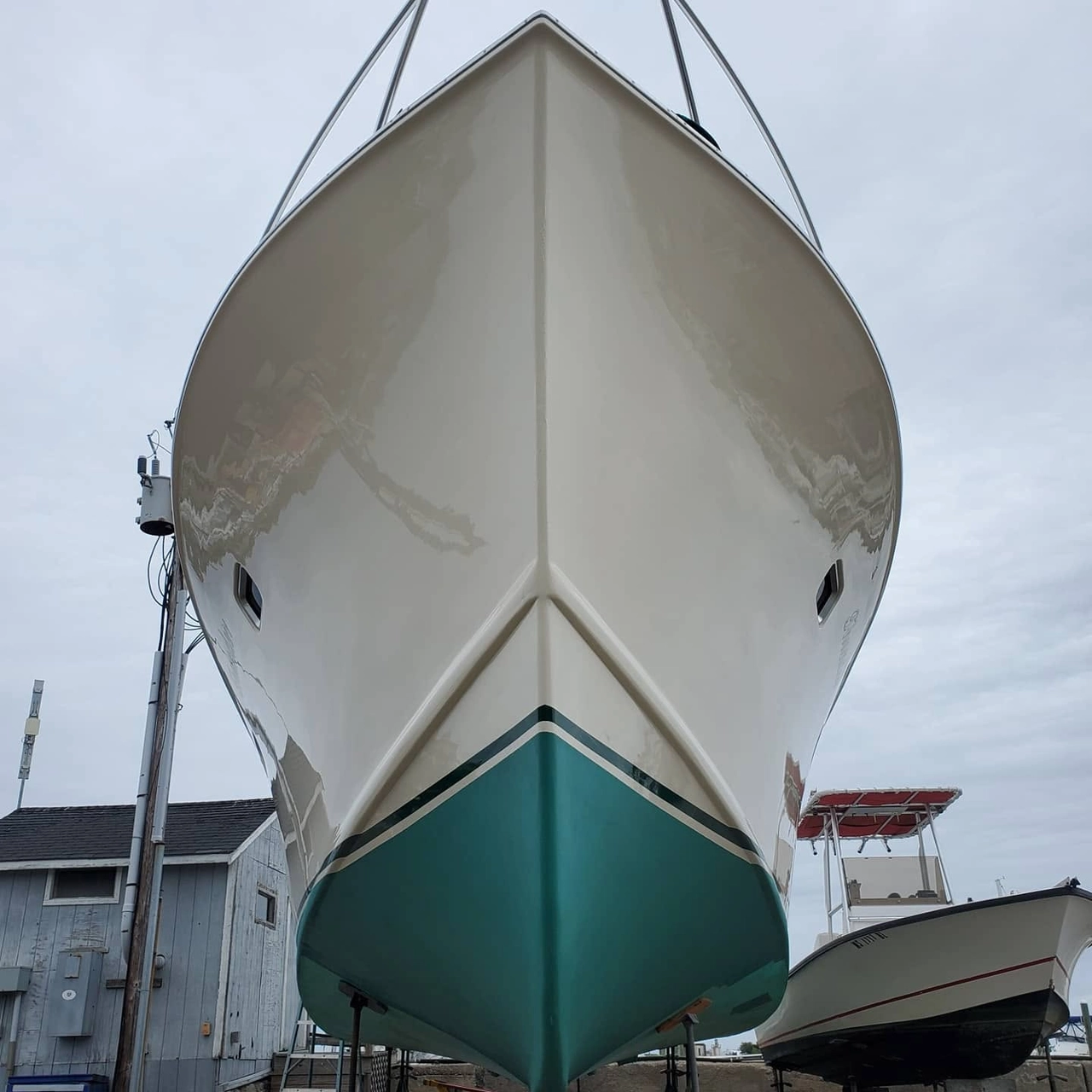 Front view of a white boat hull with teal stripe, suspended on lifting straps in a boatyard.