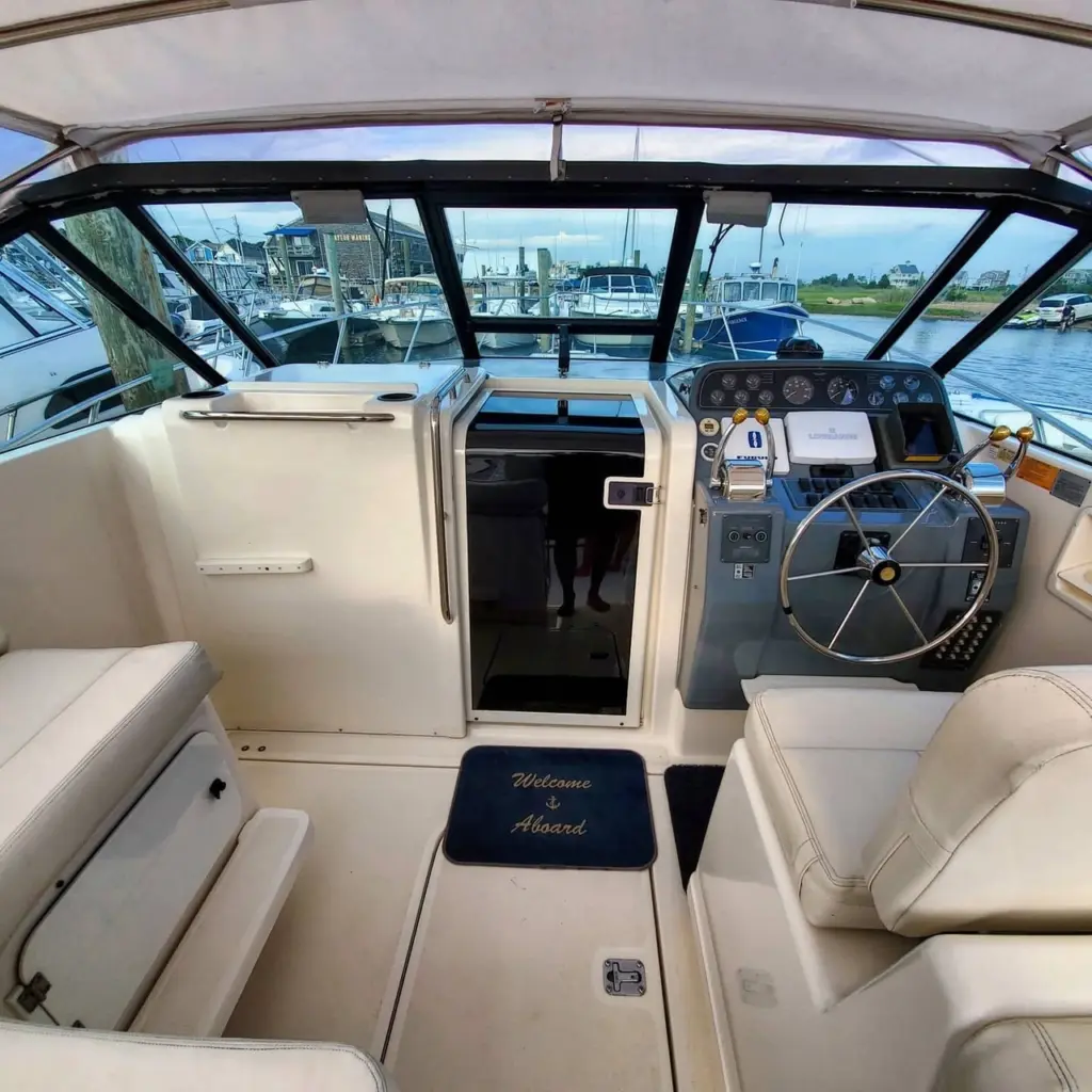 Interior view of boat cockpit with open cabin door, steering wheel, control panel, and tan seating facing marina.