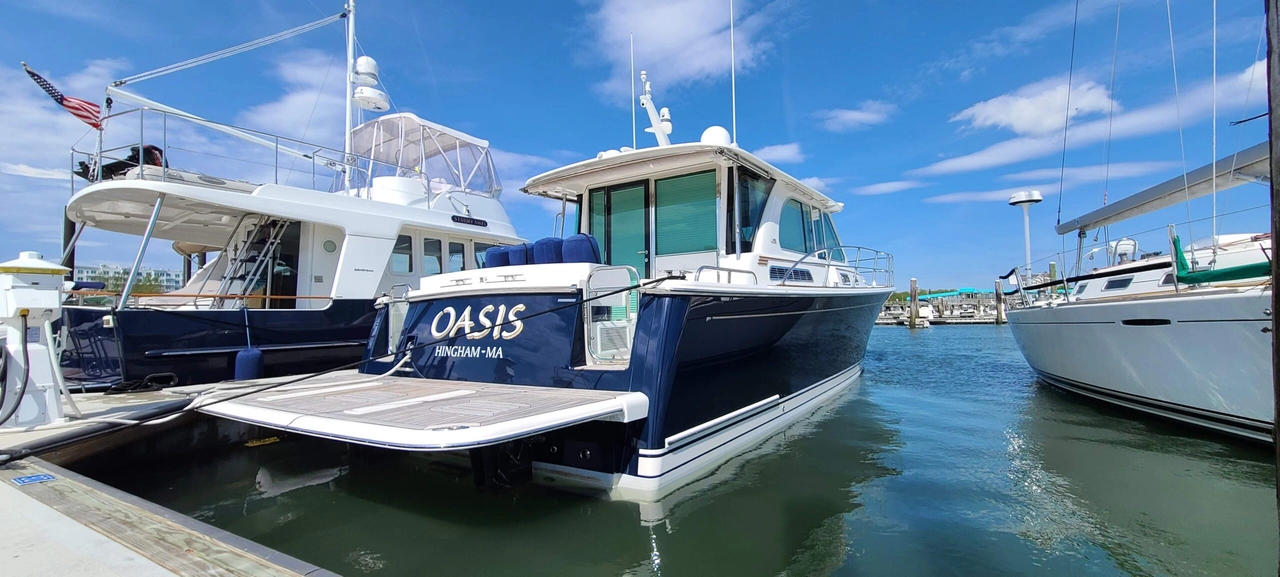 White and blue fishing boat named Oasis docked at Hingham marina with other vessels and clear blue sky.