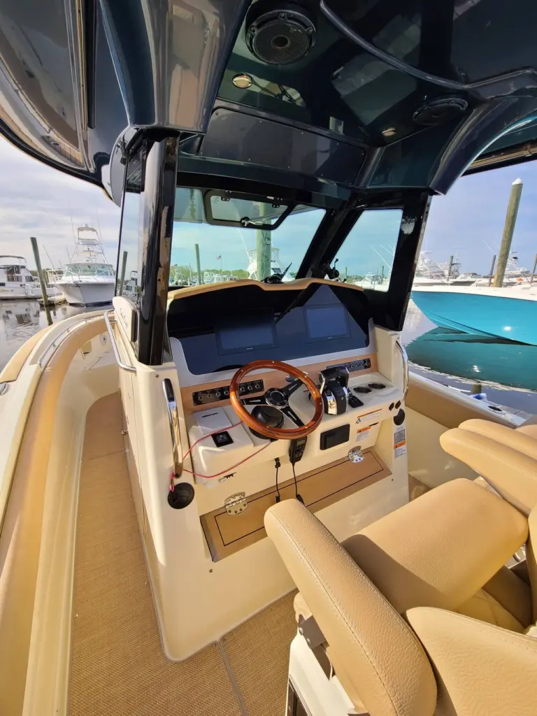 Interior view of boat helm station with wood-trimmed steering wheel, cream upholstery, and marina view through windshield.