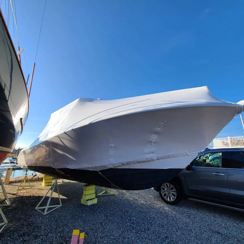 White shrink-wrapped boat on blocks in outdoor storage yard with support vehicle nearby.