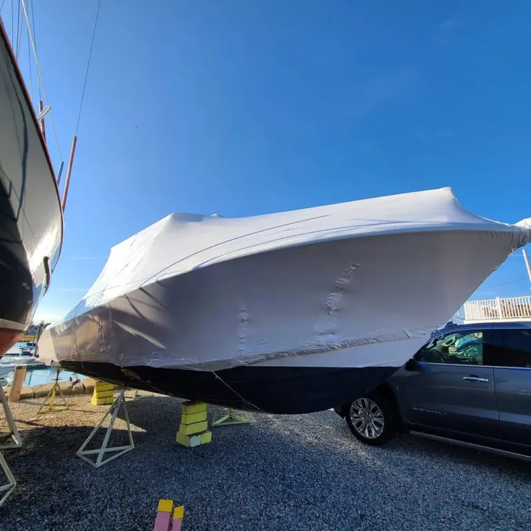 White shrink-wrapped boat on blocks in outdoor storage yard with support vehicle nearby.