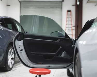 Open car door in a detailing bay with another vehicle visible and red equipment on the floor.