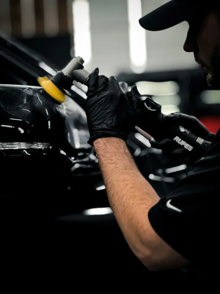 Technician in black gloves using a yellow polishing pad on a black car's surface.