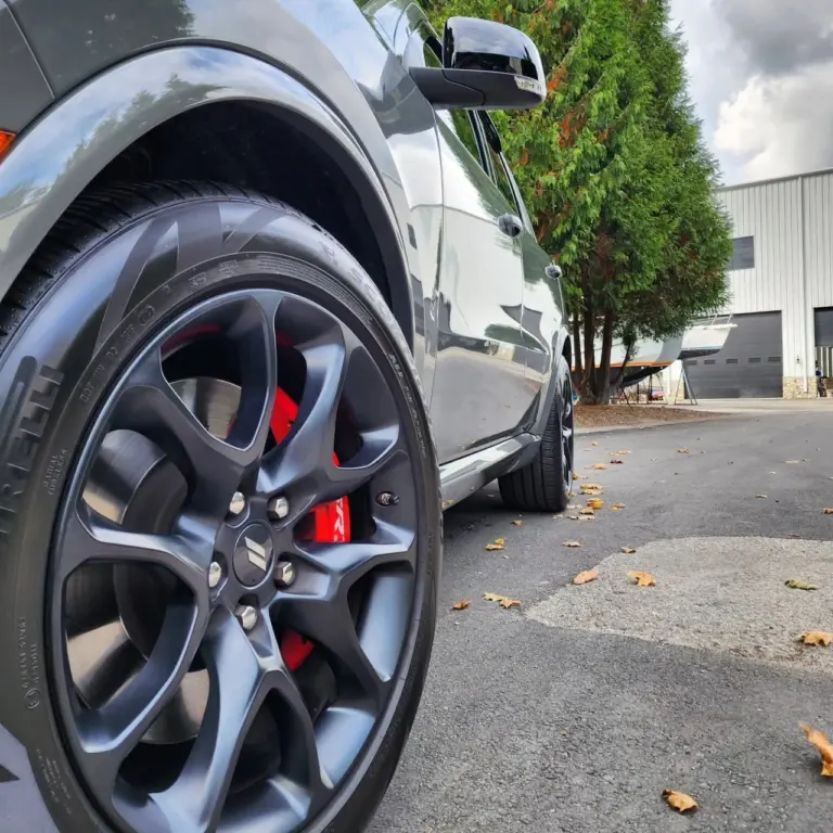Close-up of a gray sedan's custom dark wheel with red brake caliper, parked in a lot with ivy-covered building in background.