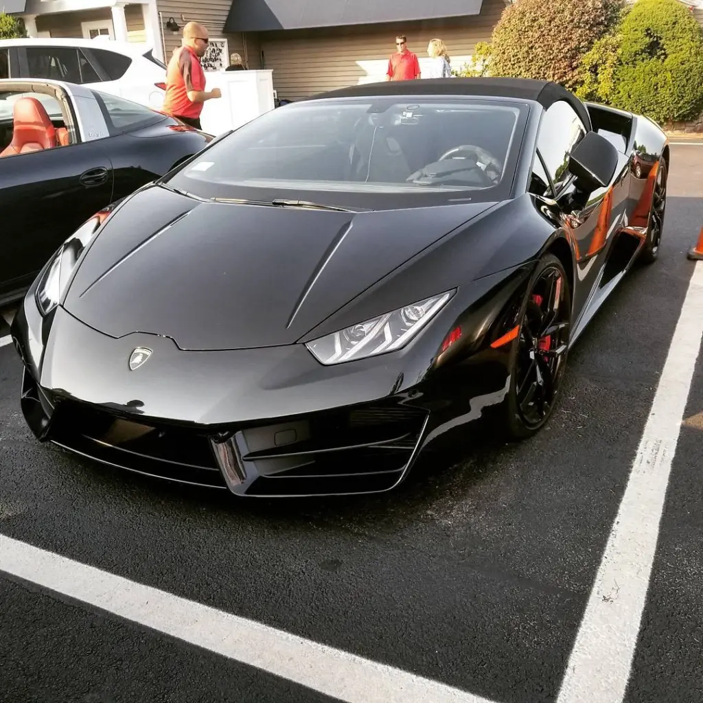 Gray and orange Lamborghini roadster with driver door open, parked in lot with staff in background.