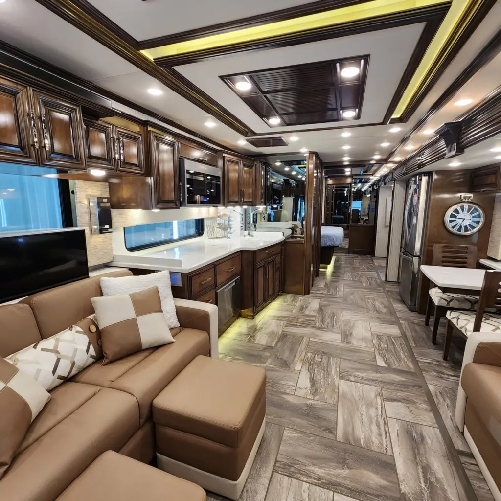 Interior of a luxury RV with tan leather seating, white pillows, wood cabinetry, and ambient lighting throughout.