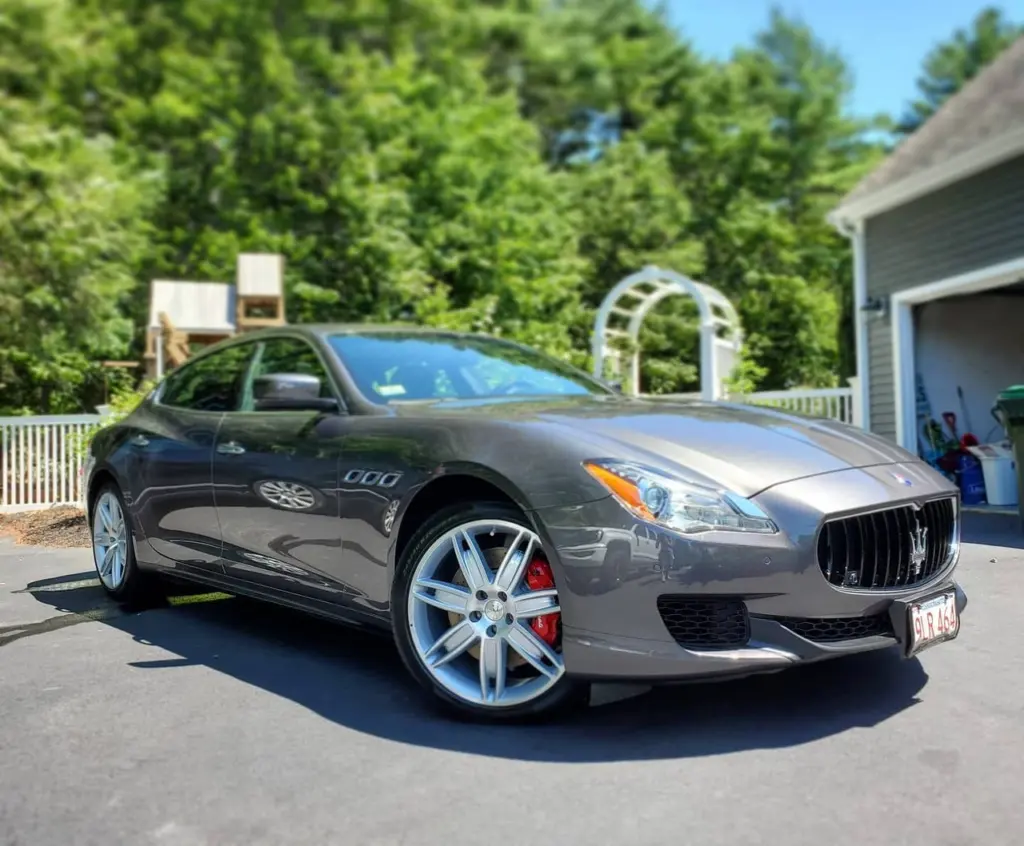 Gray Maserati Quattroporte sedan parked in driveway with garage and trees in background.