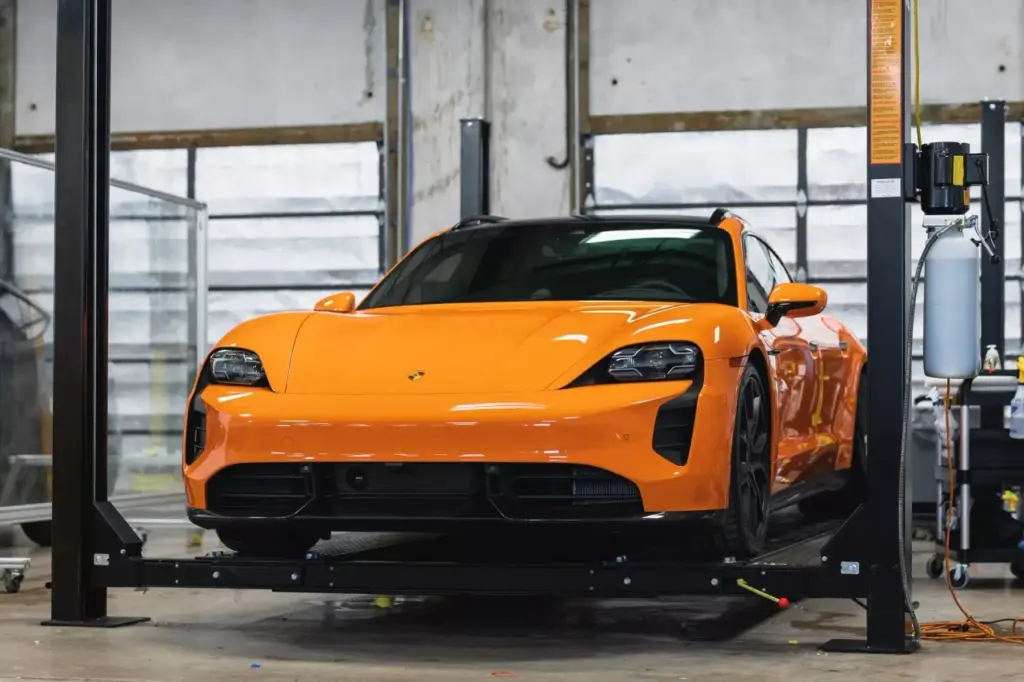 Orange Porsche sports car on a two-post lift in an indoor detailing bay with large windows.