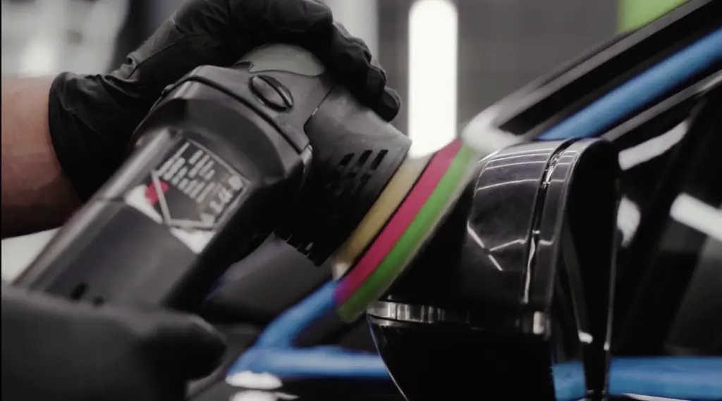 Technician in black gloves using an orbital polisher with colored foam pads on vehicle paint.