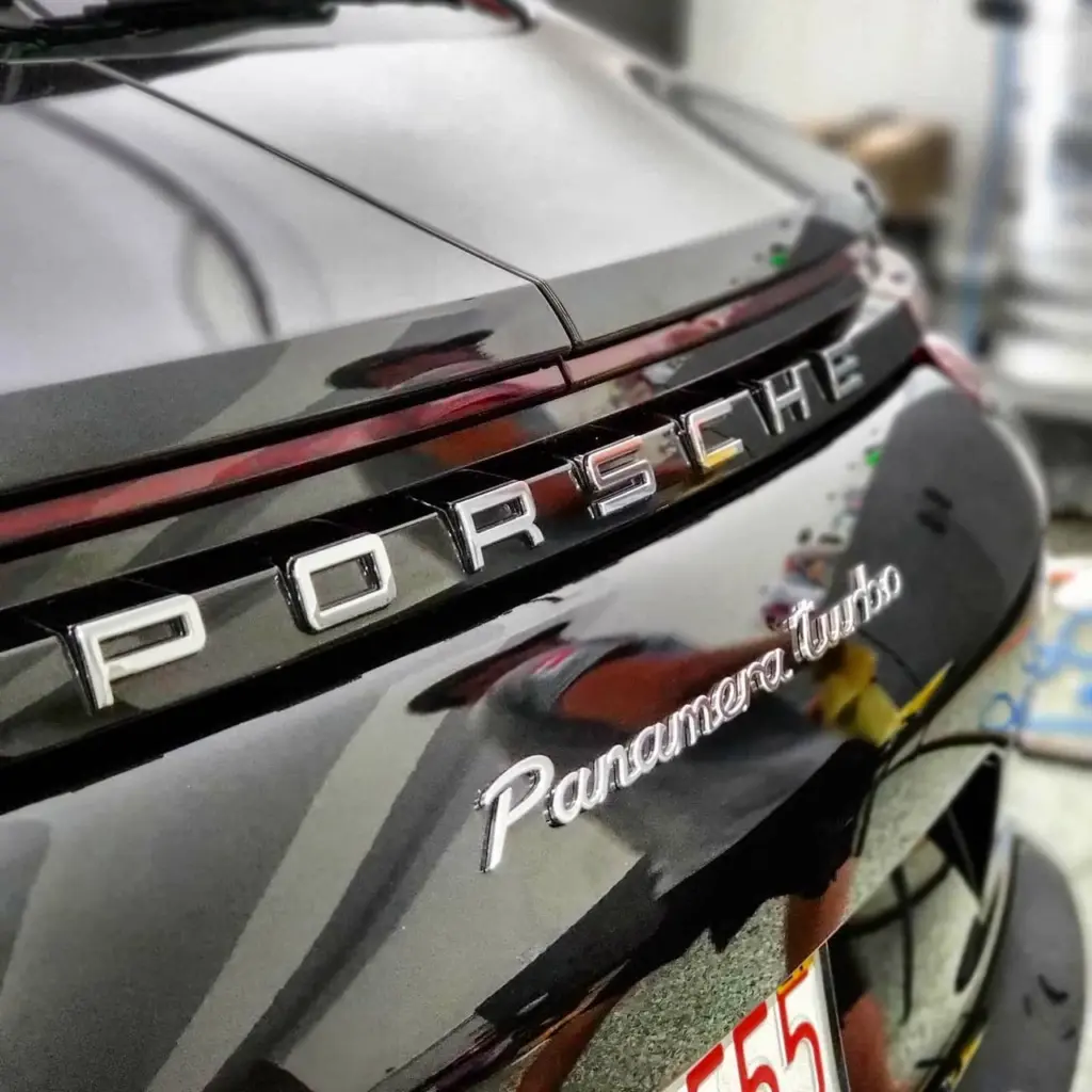 Close-up of Porsche chrome badge and red taillights on gray sedan parked at detail facility.