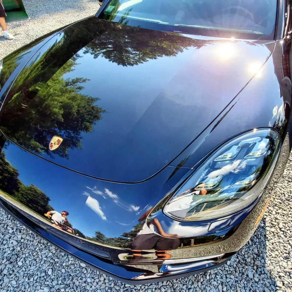 Overhead view of a dark blue Porsche hood and headlight assembly reflecting trees and sky.