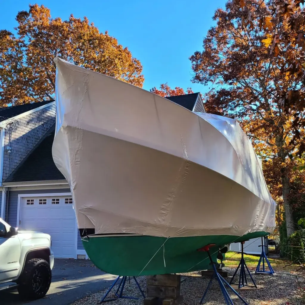 Sailboat with green hull covered in white protective tarp, supported on stands in residential driveway.