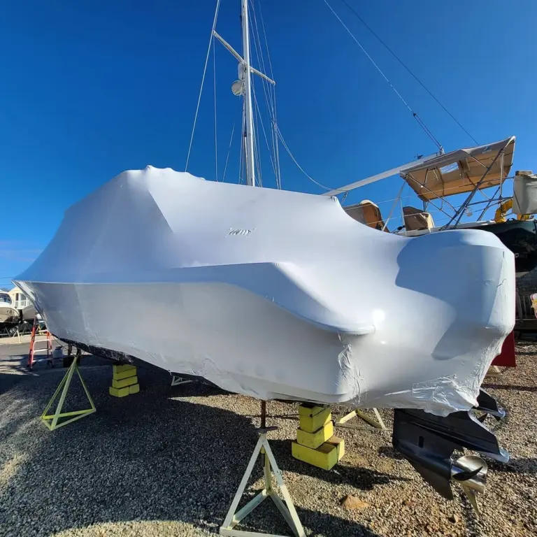White protective covering on a sailboat hull supported by yellow stands in a marina storage yard.