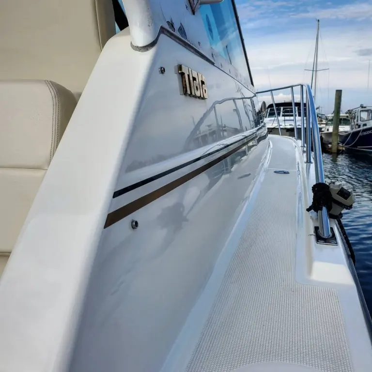 Side profile of a white boat with Tiara nameplate, showing cabin window, stripe detail, and moored sailboats in background.