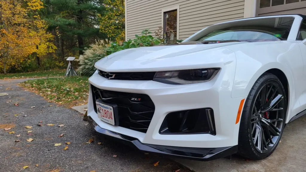 White Chevrolet Camaro ZL1 with polished front grille and headlights parked in driveway with autumn foliage.